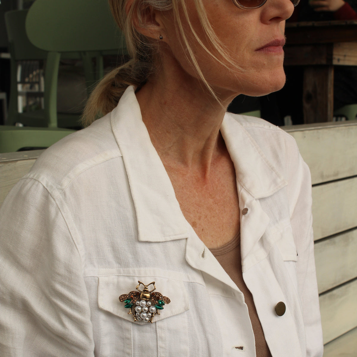 A woman wearing a white denim jacket with a decorative brooch pinned to the chest pocket, styled with sunglasses and a beige top in an outdoor setting.