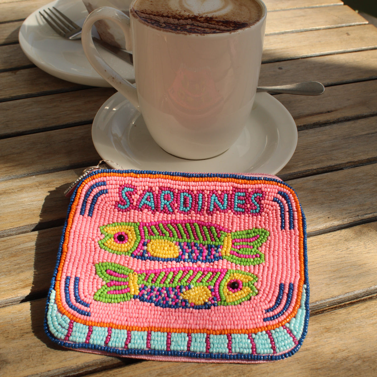 A square embroidered zip purse featuring colorful fish designs and the word 'SARDINES' in blue, placed on a wooden table next to a cup of cappuccino.