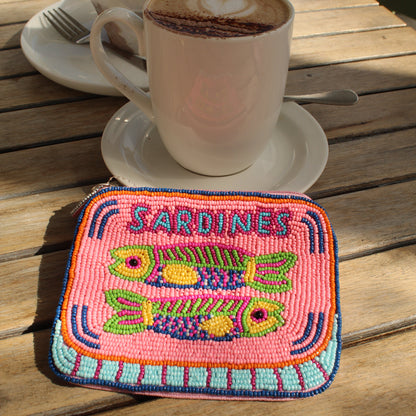 A square embroidered zip purse featuring colorful fish designs and the word 'SARDINES' in blue, placed on a wooden table next to a cup of cappuccino.
