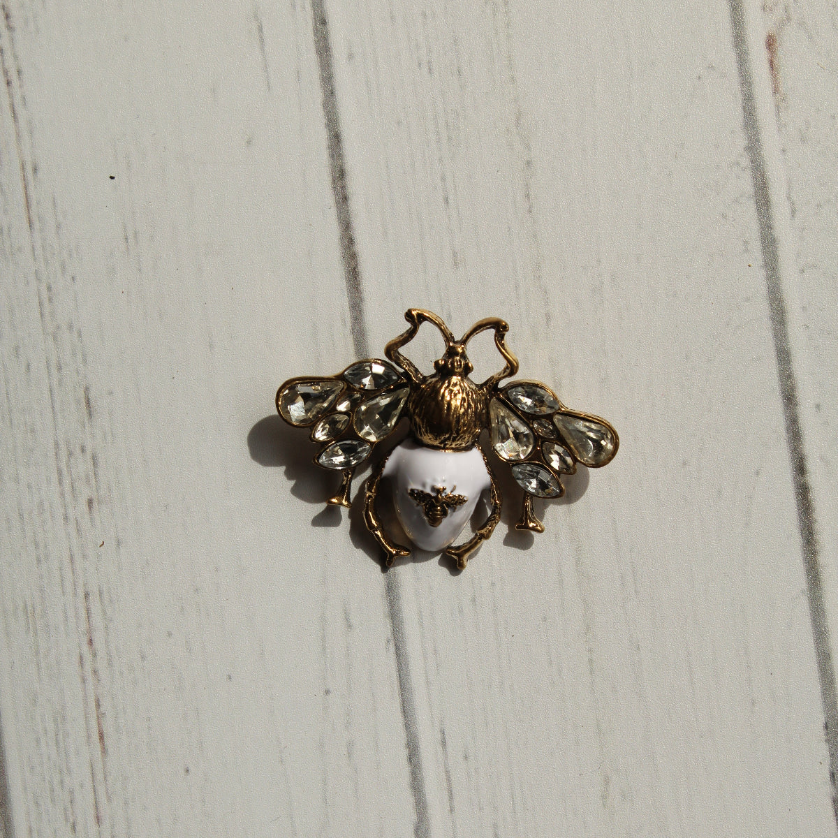 A vintage-style bee brooch with gold-tone metal, clear rhinestones, and a white enamel body, displayed on a white painted wooden surface.