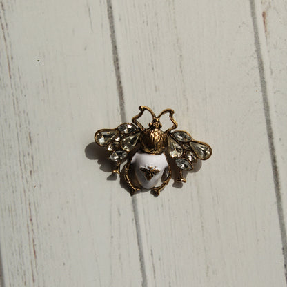 A vintage-style bee brooch with gold-tone metal, clear rhinestones, and a white enamel body, displayed on a white painted wooden surface.