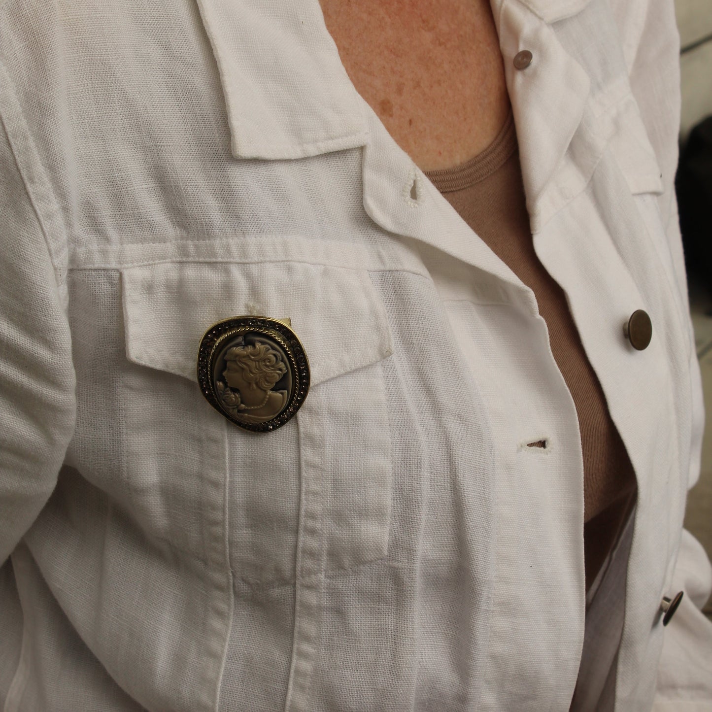A close-up of a person wearing a white linen-style button-up shirt jacket over a beige top, featuring a prominent round bronze-toned decorative pin on the chest pocket.