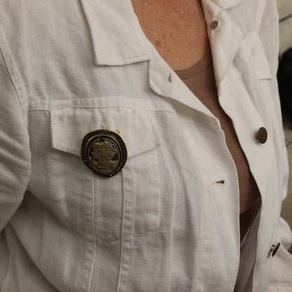 A close-up of a person wearing a white linen-style button-up shirt jacket over a beige top, featuring a prominent round bronze-toned decorative pin on the chest pocket.