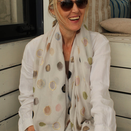 A woman wearing a white long-sleeve blouse and a white scarf with multicolored polka dots, sitting on a wooden bench in a sunlit outdoor setting.