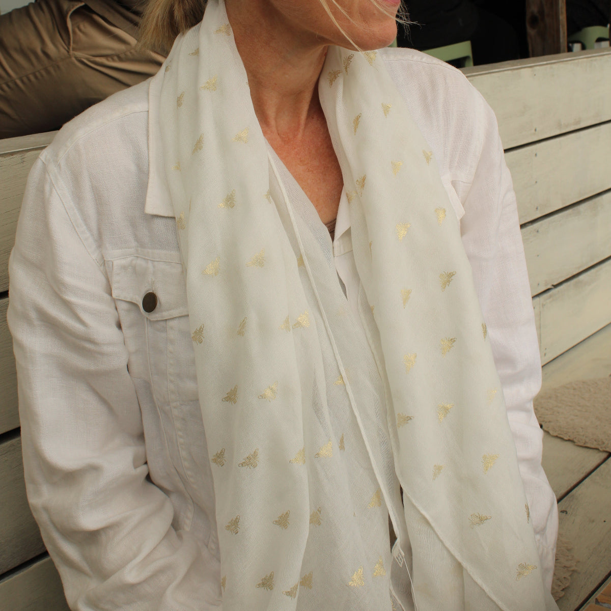A woman wearing a white button-up shirt and a white scarf with small gold bee motifs, seated on a wooden deck bench.