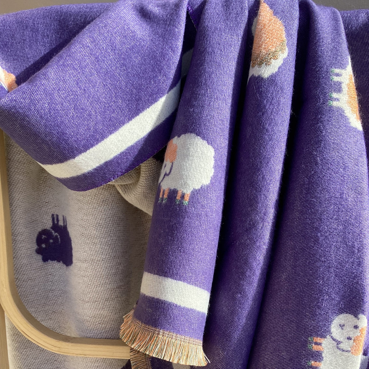 A folded purple scarf with white and orange sheep motifs and white stripe borders, draped over a wooden rack.