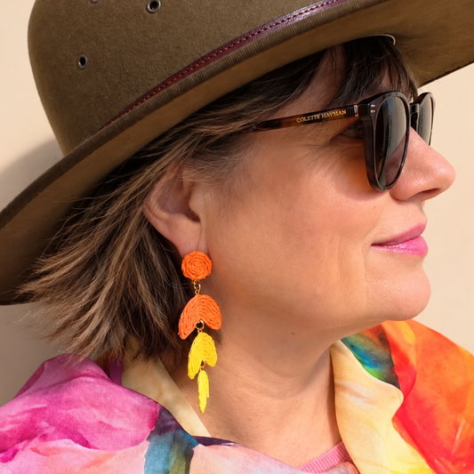A woman in profile wearing a brown wide-brim hat, dark sunglasses, and a colorful tie-dye scarf, featuring a prominent orange and yellow dangle earring.