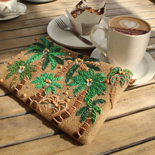 A close-up of a tan woven pouch with green palm tree embroidery and small bead details, placed on a wooden cafe table next to a cup of coffee on a saucer.