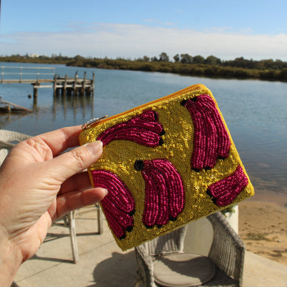 A hand holds a small zip pouch decorated with gold sequins and magenta beaded banana motifs, set against an outdoor background of a lake and dock.