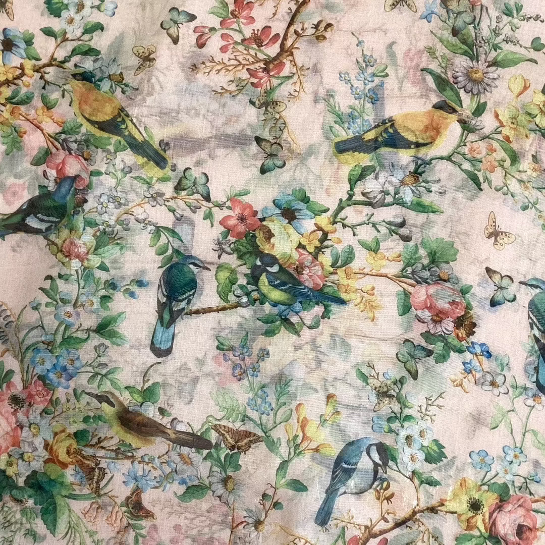 a close up view of a pale pink scarf that has blue and brown birds, butterflies and pink, blue and yellow flowers