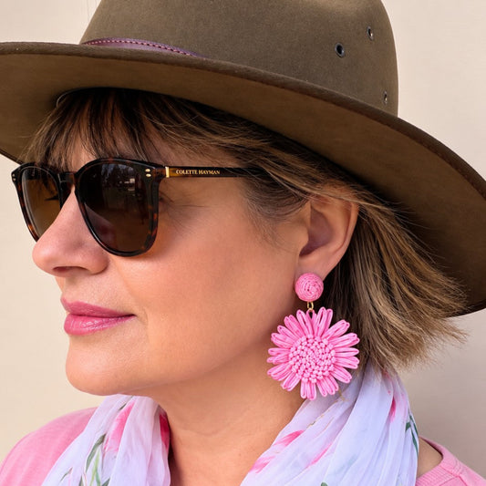 A woman in profile wearing a brown wide-brim hat, sunglasses, a pink scarf with green leaf patterns, and a pink top, featuring a large pink flower-shaped dangle earring.