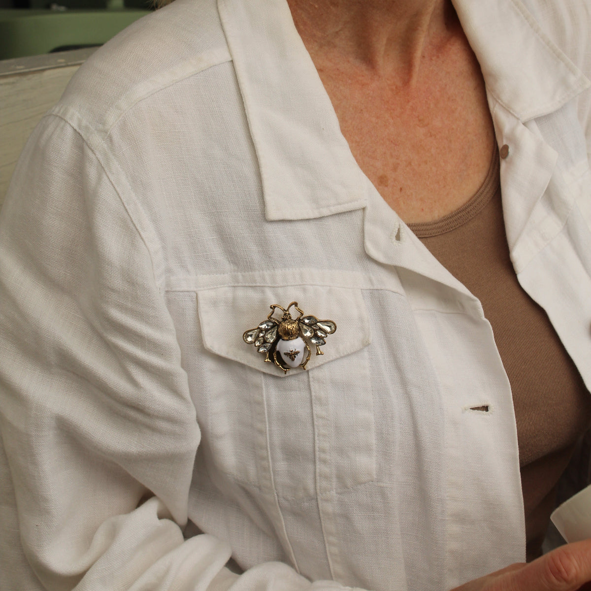 A close-up of a woman wearing a white button-up shirt over a taupe top, featuring a decorative metallic bumblebee brooch pinned to the chest pocket.
