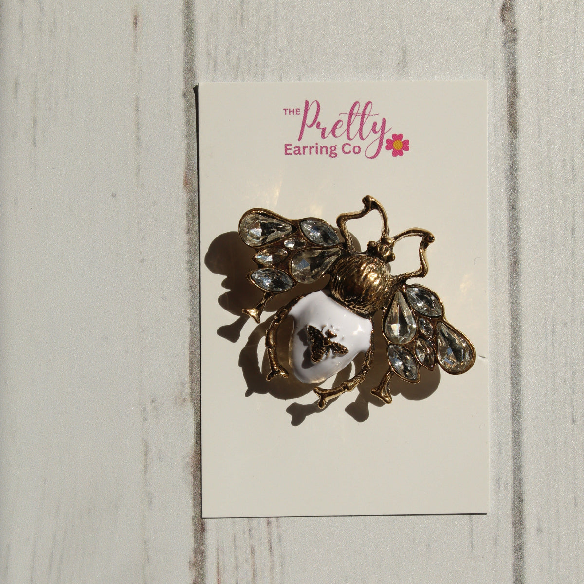 A vintage-style bee brooch with gold-tone metal, clear rhinestones, and a white enamel center, mounted on a white display card labeled 'The Pretty Earring Co' against a white wood plank background.