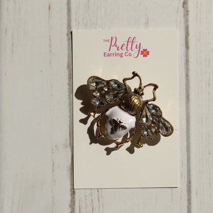 A vintage-style bee brooch with gold-tone metal, clear rhinestones, and a white enamel center, mounted on a white display card labeled 'The Pretty Earring Co' against a white wood plank background.
