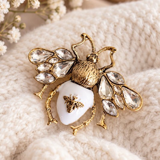 A gold-tone bee brooch with a white enamel abdomen and clear rhinestone wings, displayed on a soft cream knit fabric with small white flowers in the background.