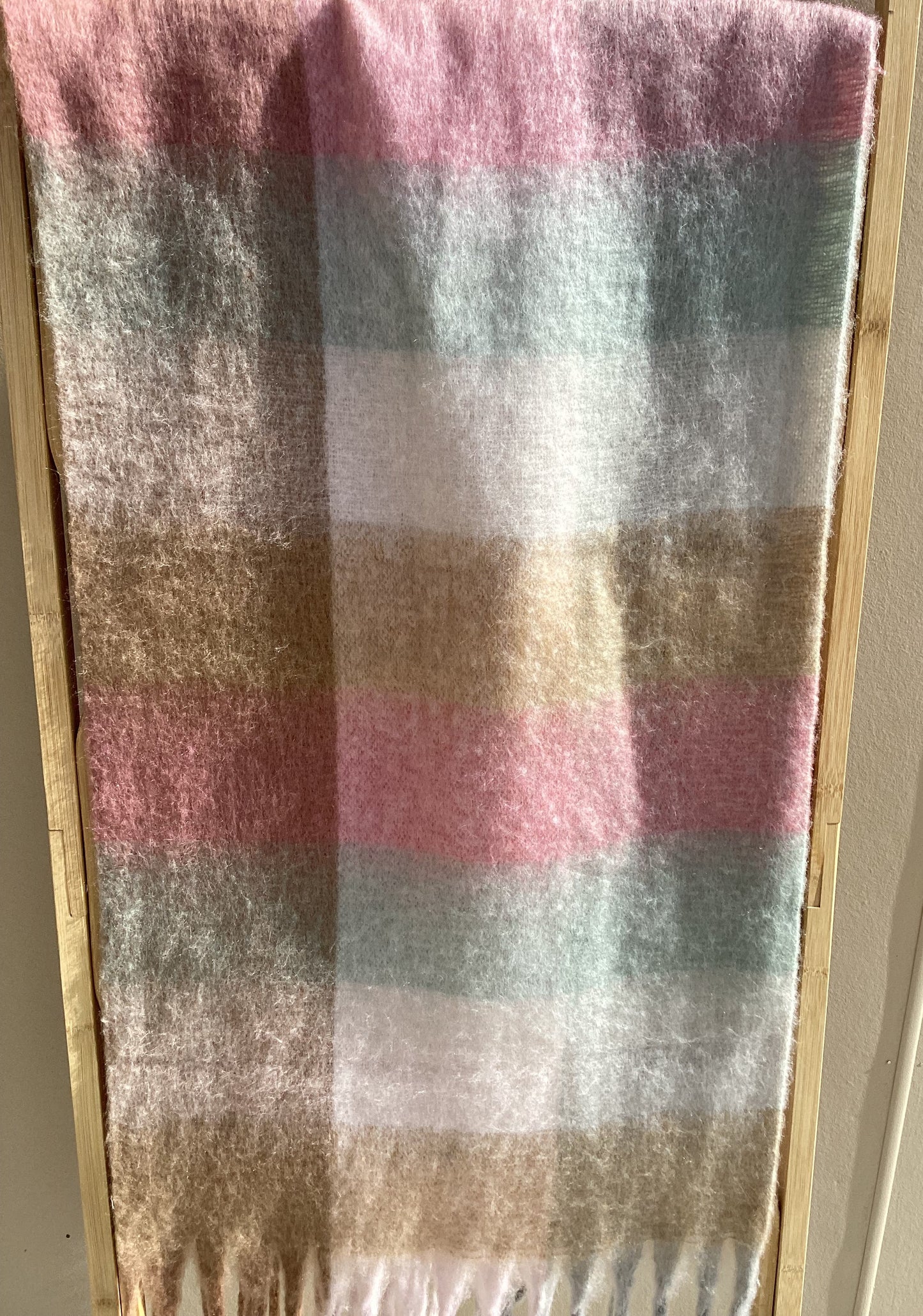 A soft winter scarf with fringe on the edges, displayed flat on a light wood surface against a beige wall.