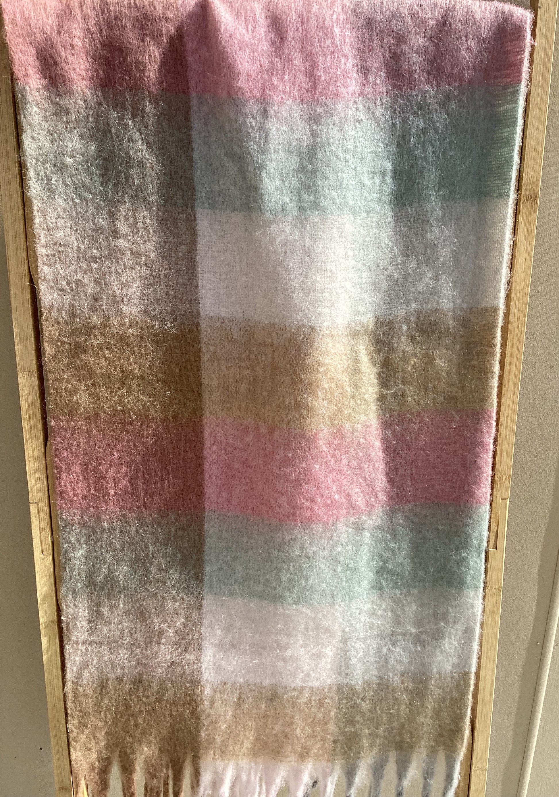A soft winter scarf with fringe on the edges, displayed flat on a light wood surface against a beige wall.