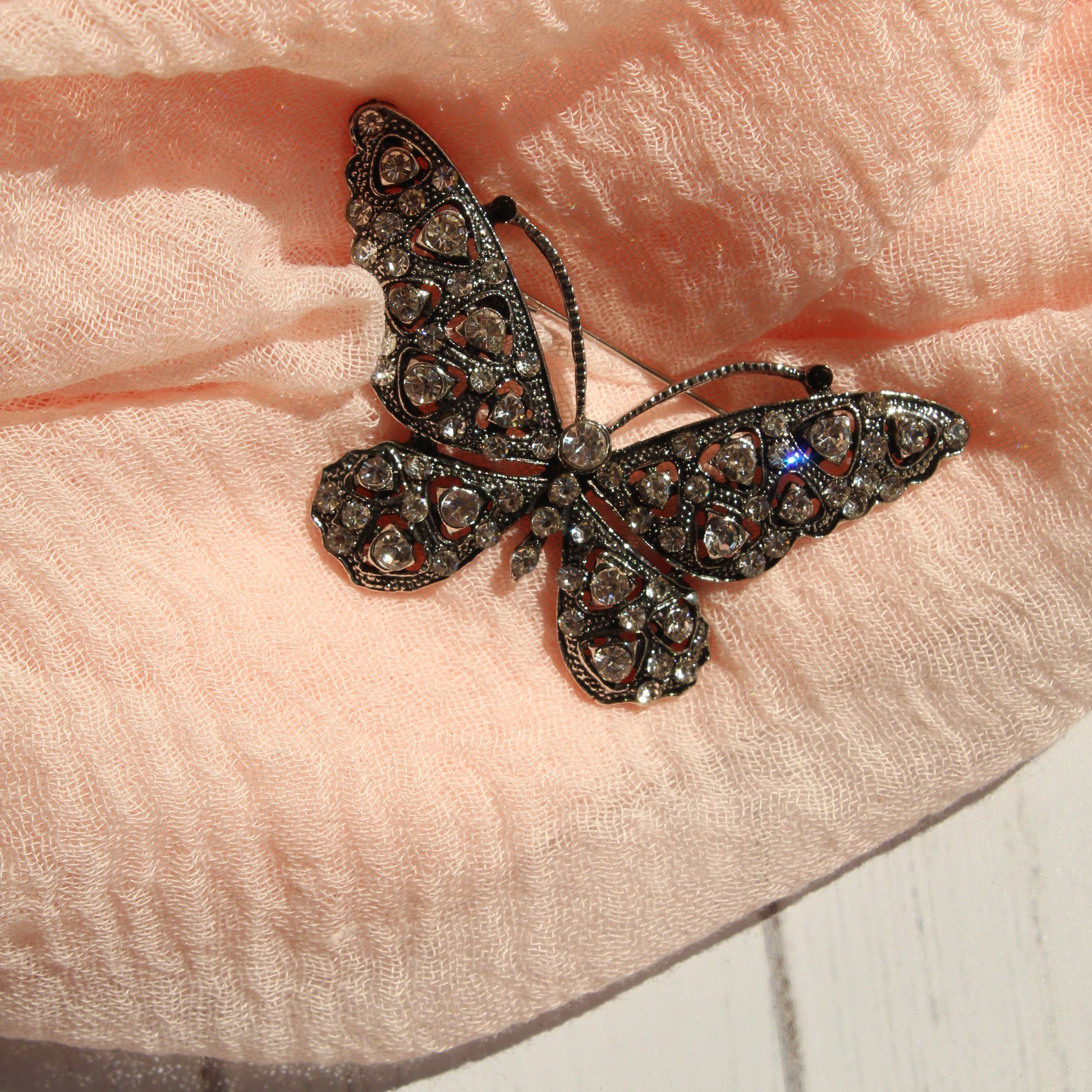 A butterfly-shaped brooch with dark filigree and sparkling clear rhinestones, resting on a soft peach-colored textured scarf