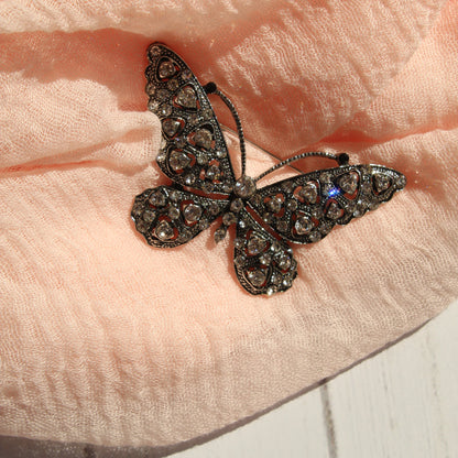 A butterfly-shaped brooch with dark filigree and sparkling clear rhinestones, resting on a soft peach-colored textured scarf