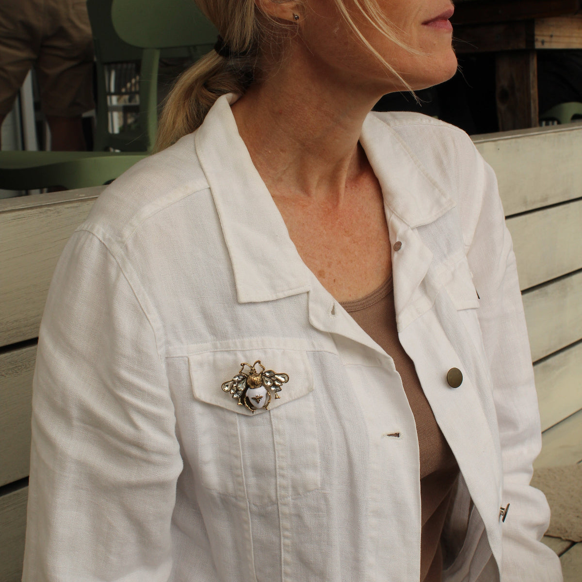 A woman in a white denim jacket with a decorative brooch on the chest pocket, wearing sunglasses and a tan top, photographed in an outdoor setting.