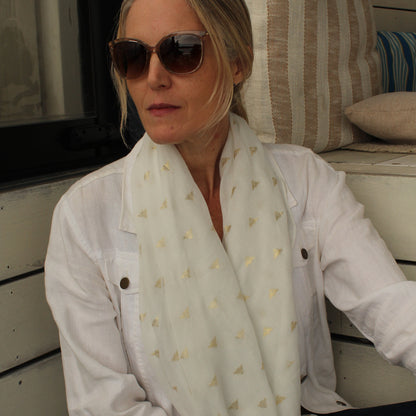 A woman wearing sunglasses, a white button-up shirt, and a white scarf with small gold bee pattern, sitting in front of a white wooden wall with cushions.