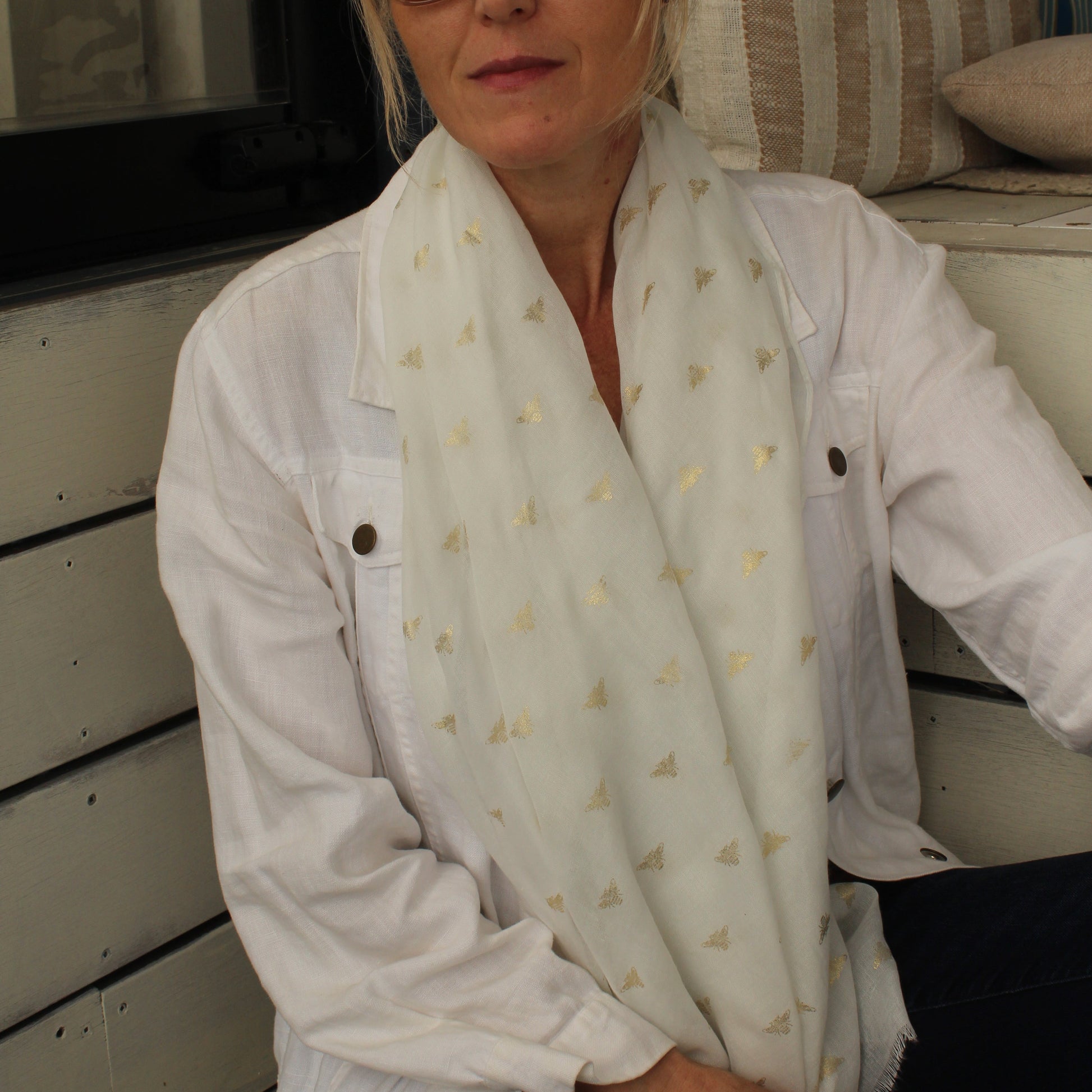 A woman wearing a white button-up shirt and a white scarf with small gold bees, sitting in front of a white wooden wall.