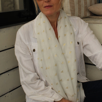 A woman wearing a white button-up shirt and a white scarf with small gold bees, sitting in front of a white wooden wall.