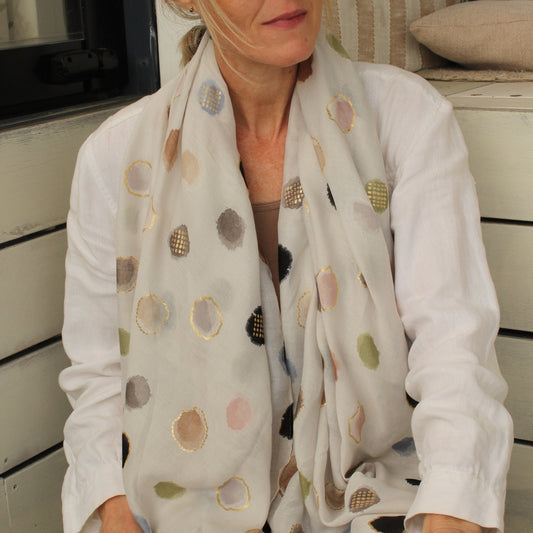 A woman wearing a white long-sleeve shirt and dark sunglasses, draped in a white scarf with multicolored polka dots, sitting on a wooden bench.