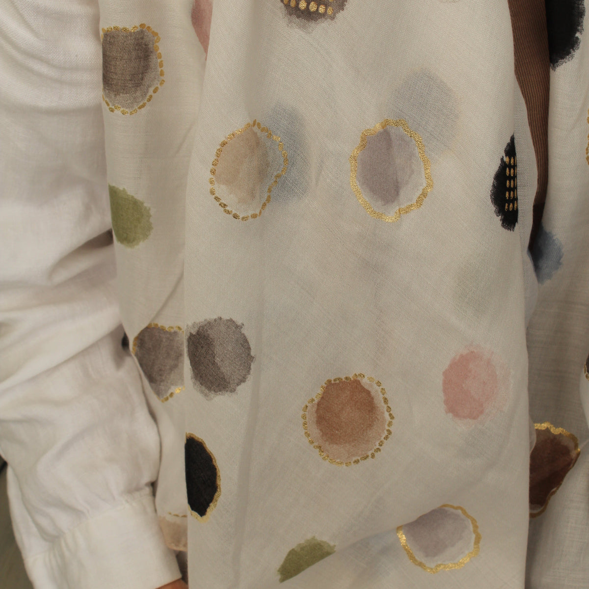 A close-up of a white or off-white scarf with scattered abstract watercolor-style polka dots in muted gray, brown, pink, green, and black, each outlined with metallic gold detailing. A person wearing a white long-sleeve top is partially visible at the left edge.
