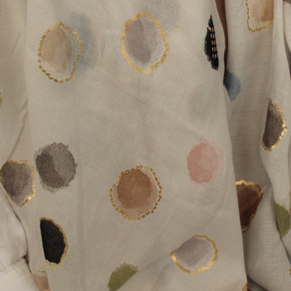 A close-up of a white or off-white fabric with scattered abstract watercolor-style polka dots in muted gray, brown, pink, black, and olive green, each outlined with metallic gold trim.