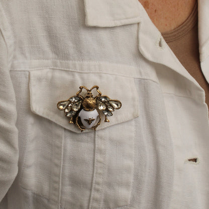 A close-up of a woman wearing a white linen button-up shirt, featuring a decorative gold and silver bee brooch pinned to the chest pocket.