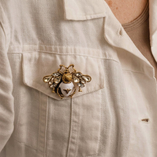 A close-up of a woman wearing a white linen button-up shirt, featuring a decorative gold and silver bee brooch pinned to the chest pocket.