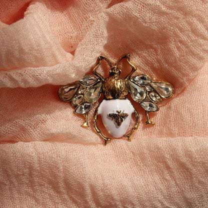 A gold-tone bumblebee brooch with a white enamel center and clear rhinestone wings, resting on draped peach-colored scarf