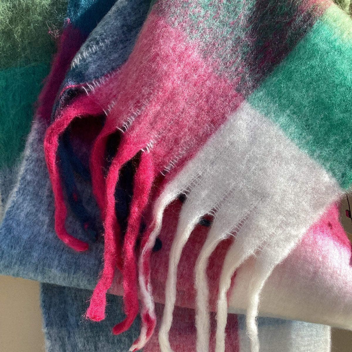 A close-up of a soft woven scarf with a plaid pattern in teal green, pink, white, and blue, featuring white fringe tassels.