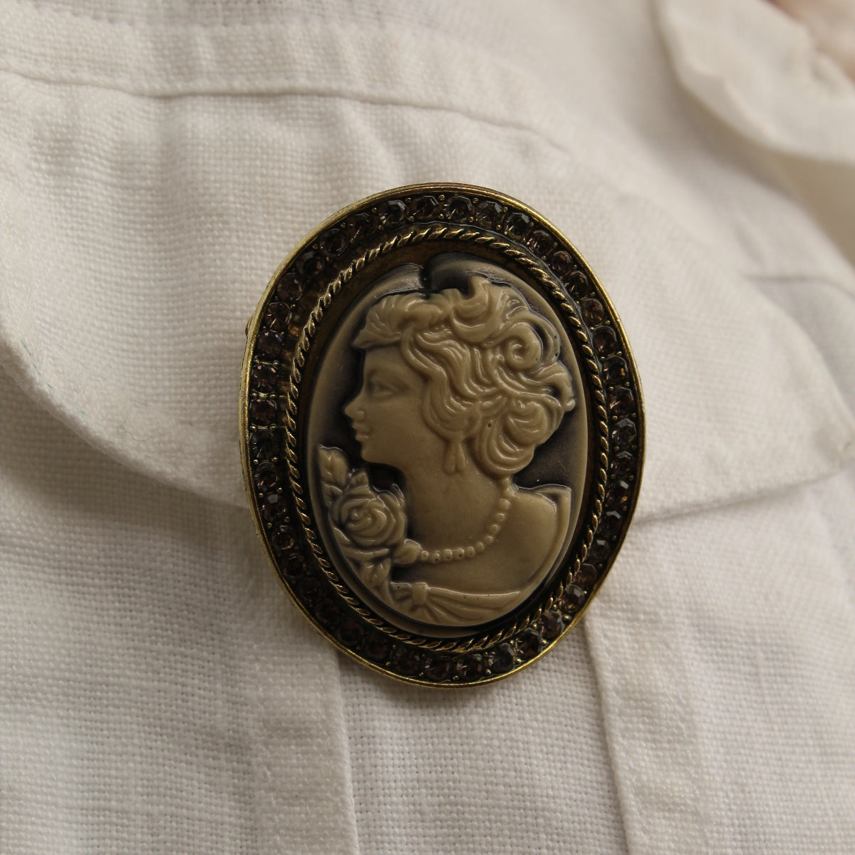An oval vintage-style cameo brooch with a dark ornate frame and a profile portrait in warm beige tones, pinned onto a white textured fabric.