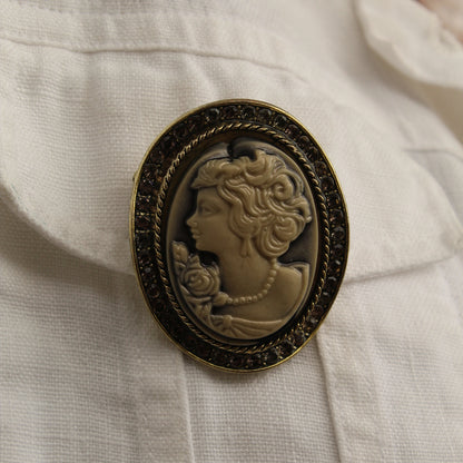 An oval vintage-style cameo brooch with a dark ornate frame and a profile portrait in warm beige tones, pinned onto a white textured fabric.