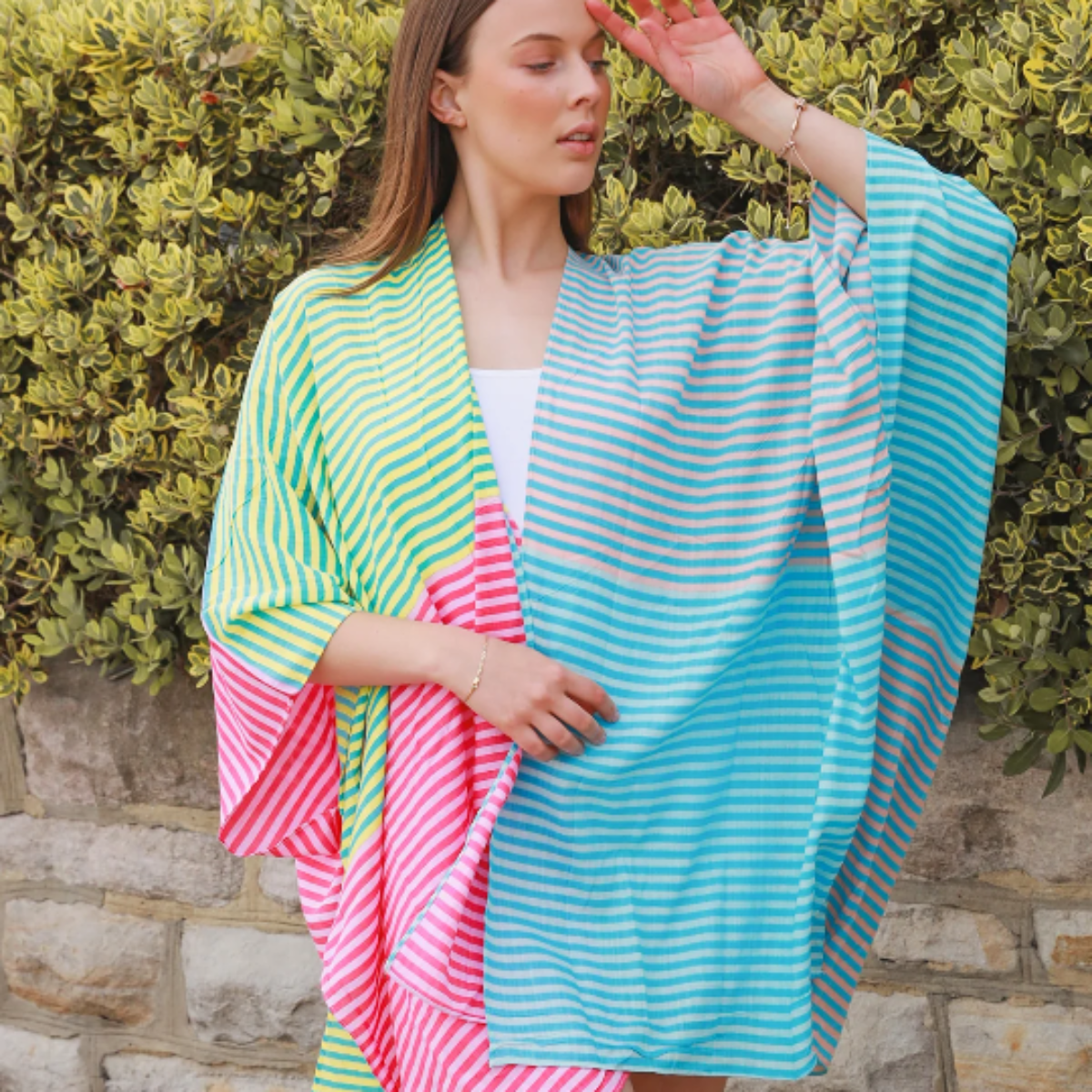 Woman holding a colorful striped kimono against a stone wall and green hedge background