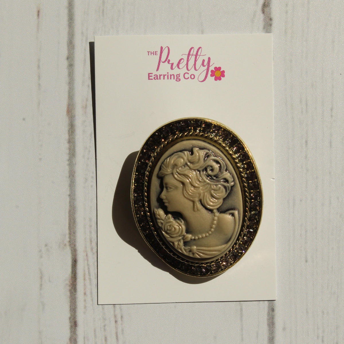 An oval vintage-style cameo pin with a gold-toned border and cream-colored profile portrait, mounted on a white display card labeled 'The Pretty Earring Co' and photographed against a white wood plank background.