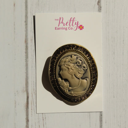 An oval vintage-style cameo pin with a gold-toned border and cream-colored profile portrait, mounted on a white display card labeled 'The Pretty Earring Co' and photographed against a white wood plank background.